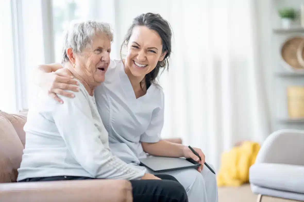 A caregiver hugs a smiling older woman on a couch.