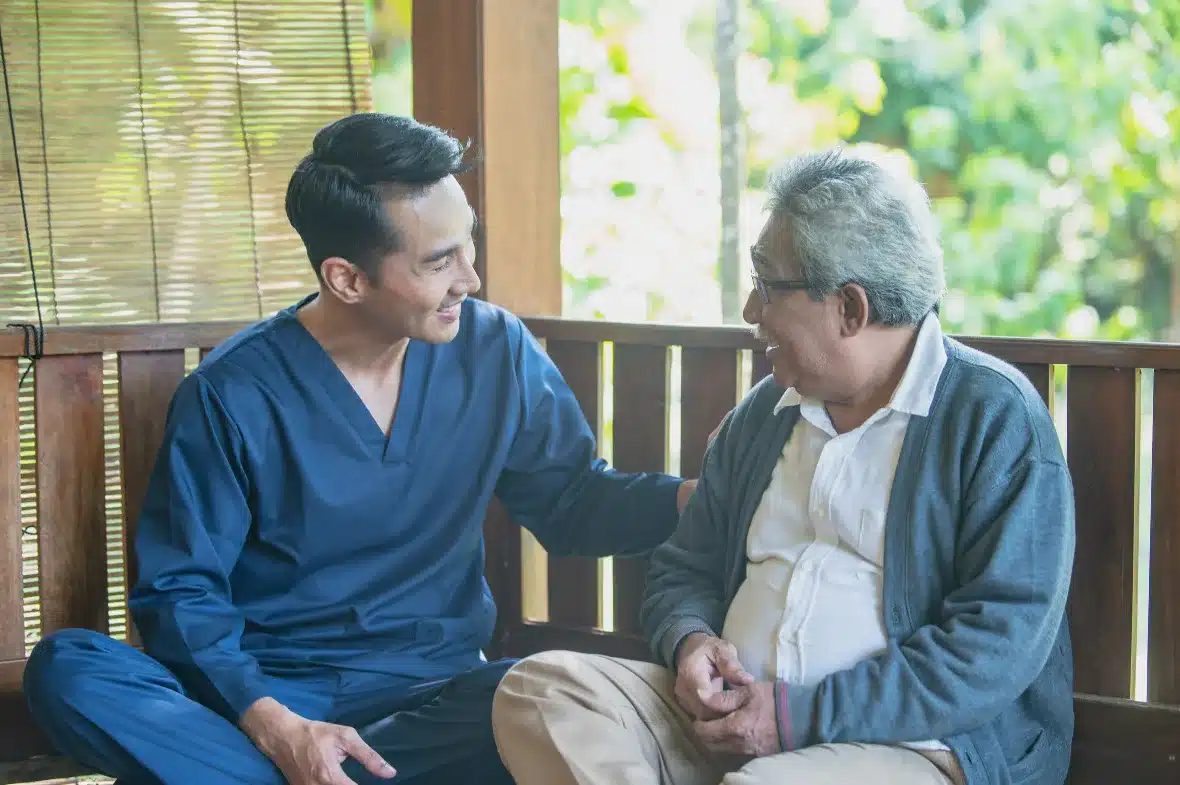 A caregiver and an older man talk on a porch bench.