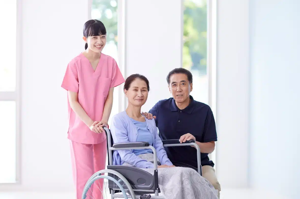 A smiling caregiver in pink scrubs stands beside a woman in a wheelchair, accompanied by a man. The setting is bright, conveying a caring and supportive atmosphere.
