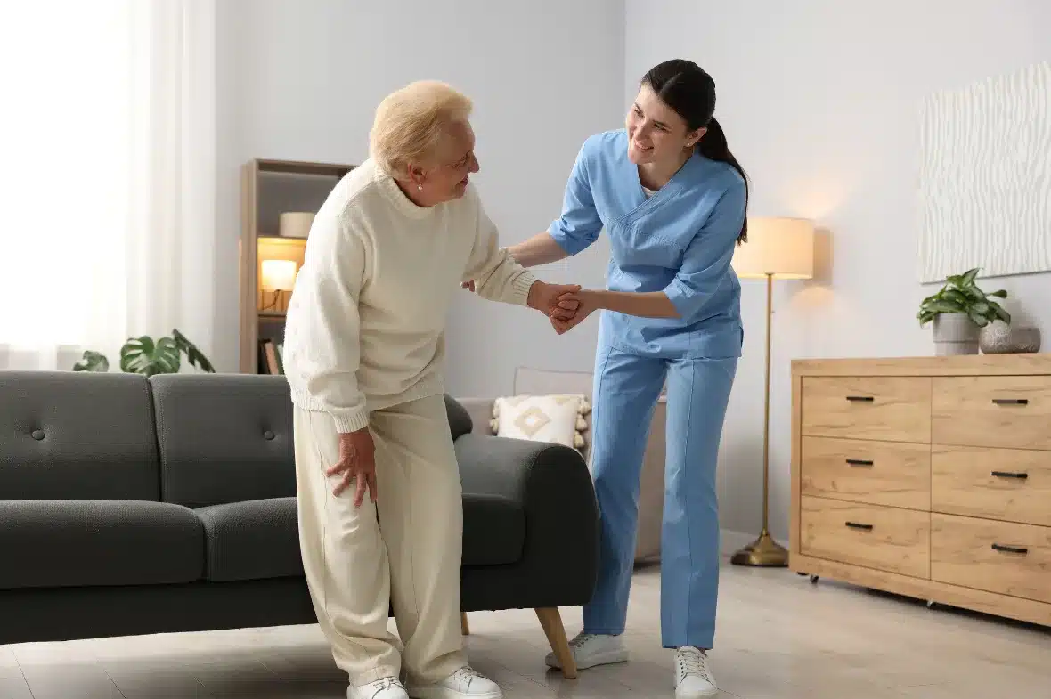 A caregiver hands a cup to a smiling older woman seated in an armchair.