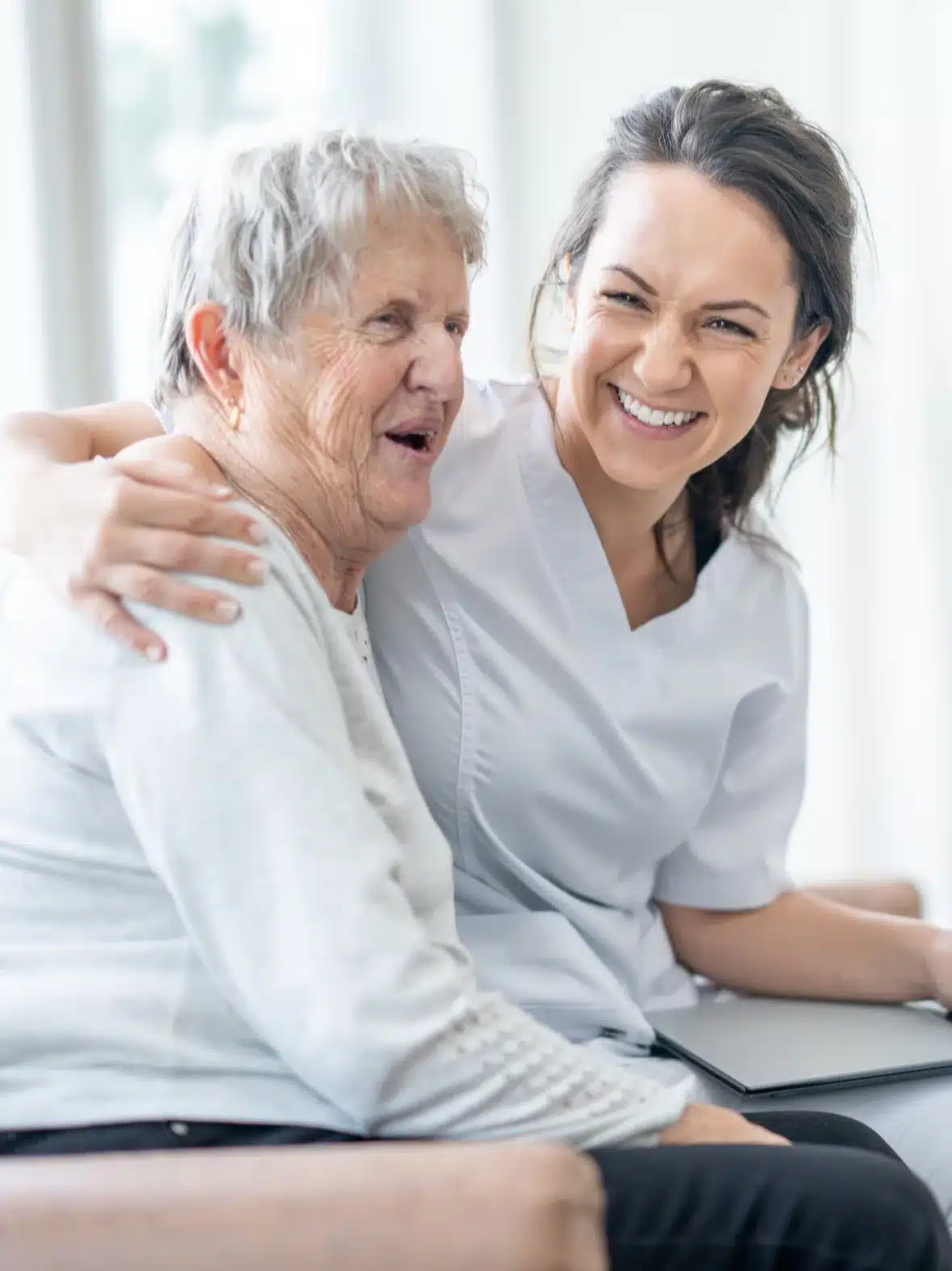 A smiling caregiver hugs an older woman on a sofa.