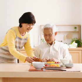 An elderly man sits at a kitchen table with breakfast. A woman stands beside him, smiling, wearing an apron. The scene is warm and homey.