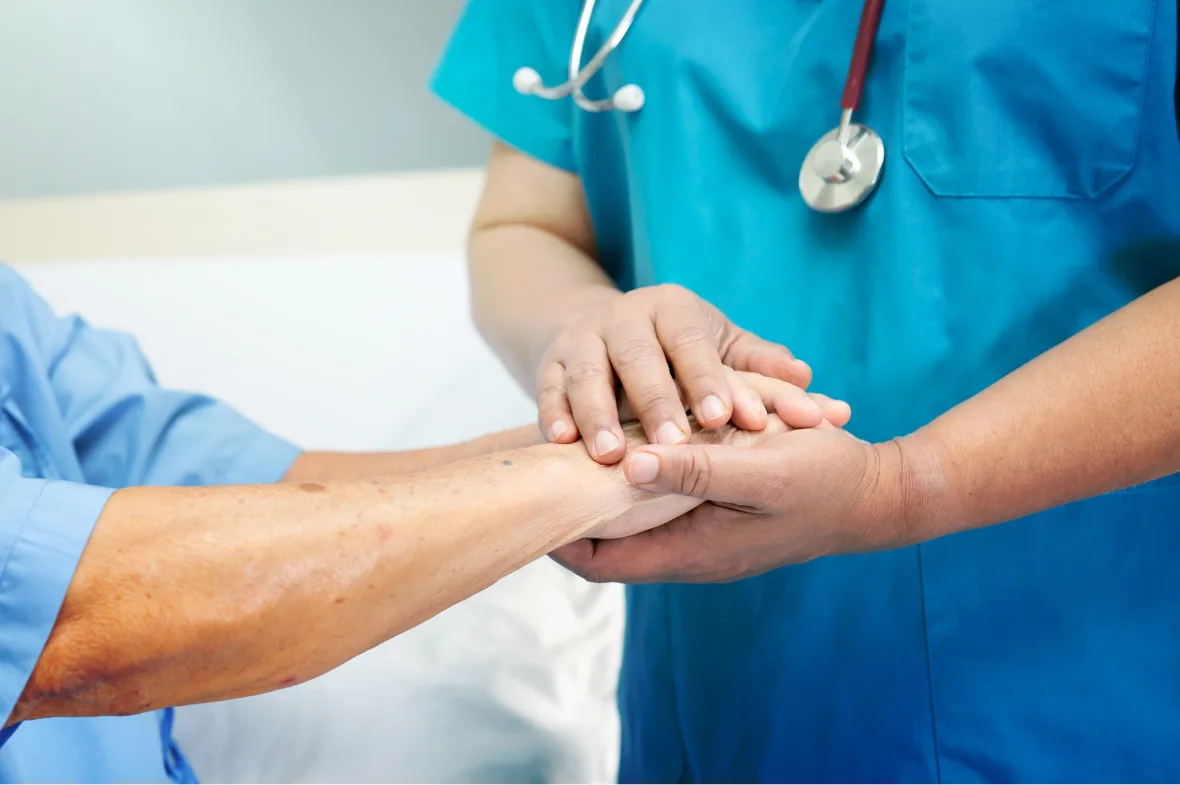 Healthcare professional in blue scrubs and stethoscope gently holding an elderly person's hands, conveying care and compassion in a clinical setting.