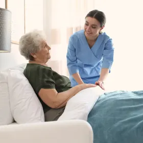 A caregiver in a blue uniform gently attends to an elderly woman in bed, creating a warm atmosphere of care and support in a bright room.