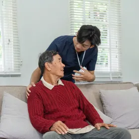 A caregiver in blue scrubs gently supports an elderly man in a red sweater sitting on a couch, conveying care and attentiveness in a well-lit room.