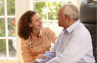 A caregiver smiles warmly at an older man, sitting by a window. The setting is bright and cozy, conveying care and companionship.