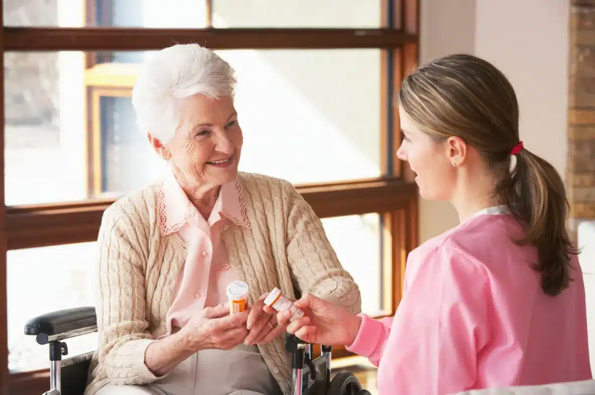 An elderly woman in a wheelchair smiles while receiving medication from a nurse in pink scrubs. Bright, warm indoor setting with large windows.