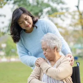 A smiling nurse in teal scrubs stands with a stethoscope, gently holding the shoulders of a seated elderly woman in a gray sweater, conveying warmth and care.