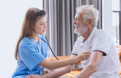 A nurse in blue scrubs is smiling while using a stethoscope on an elderly man in a white polo. They sit in a well-lit room, conveying warmth and care.