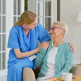 A caregiver in blue scrubs sits beside a smiling elderly woman, their hands gently on each other's shoulders, conveying warmth and compassion in a home setting.