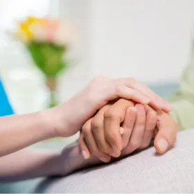 Two hands gently holding another hand, conveying comfort and support. A blurred bouquet of flowers in the background adds a warm tone.