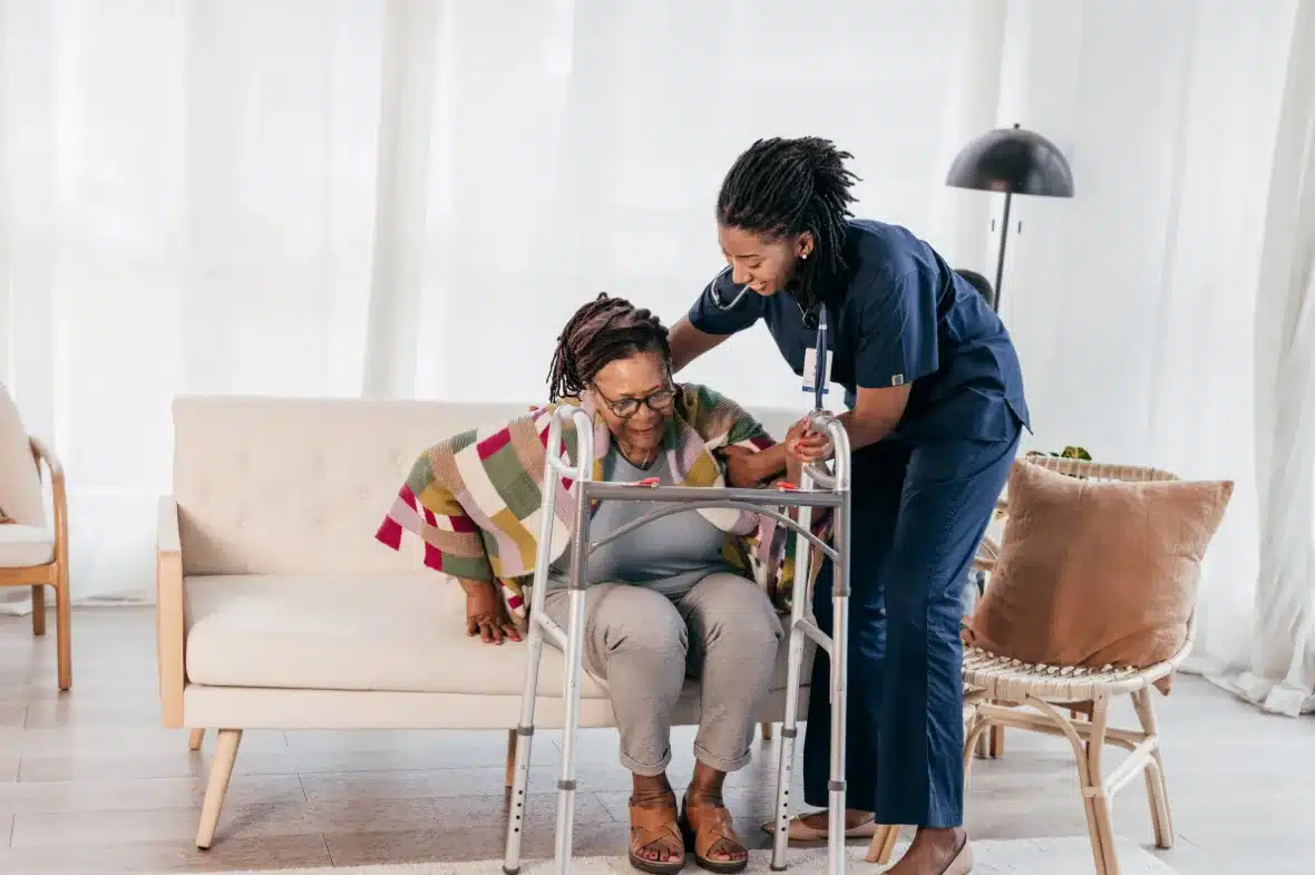 A nurse in blue scrubs assists an elderly woman with a walker. The woman, draped in a colorful blanket, is sitting on a sofa, showing a comforting scene.