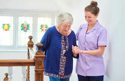 A caregiver in a lavender uniform assists an elderly woman walking near a staircase. The setting is bright, with stained glass windows in the background.