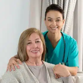 A smiling nurse in teal scrubs stands with a stethoscope, gently holding the shoulders of a seated elderly woman in a gray sweater, conveying warmth and care.