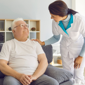 An elderly woman comforts a man sitting on a sofa. She holds his hand and looks concerned, creating a supportive and caring atmosphere in a cozy room.