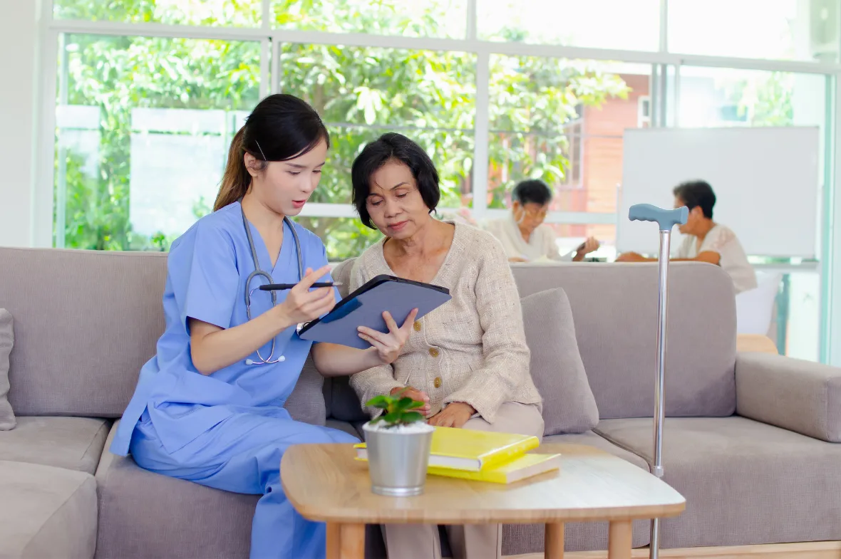 A nurse in blue scrubs shows an elderly woman something on a tablet while sitting together on a couch. A crutch leans beside them, and other seniors are interacting in the background in a bright, spacious room.