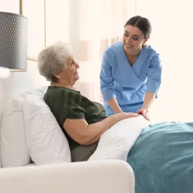 A smiling caregiver in blue scrubs attentively tends to an elderly woman sitting in bed. The room is bright and serene, conveying warmth and care.
