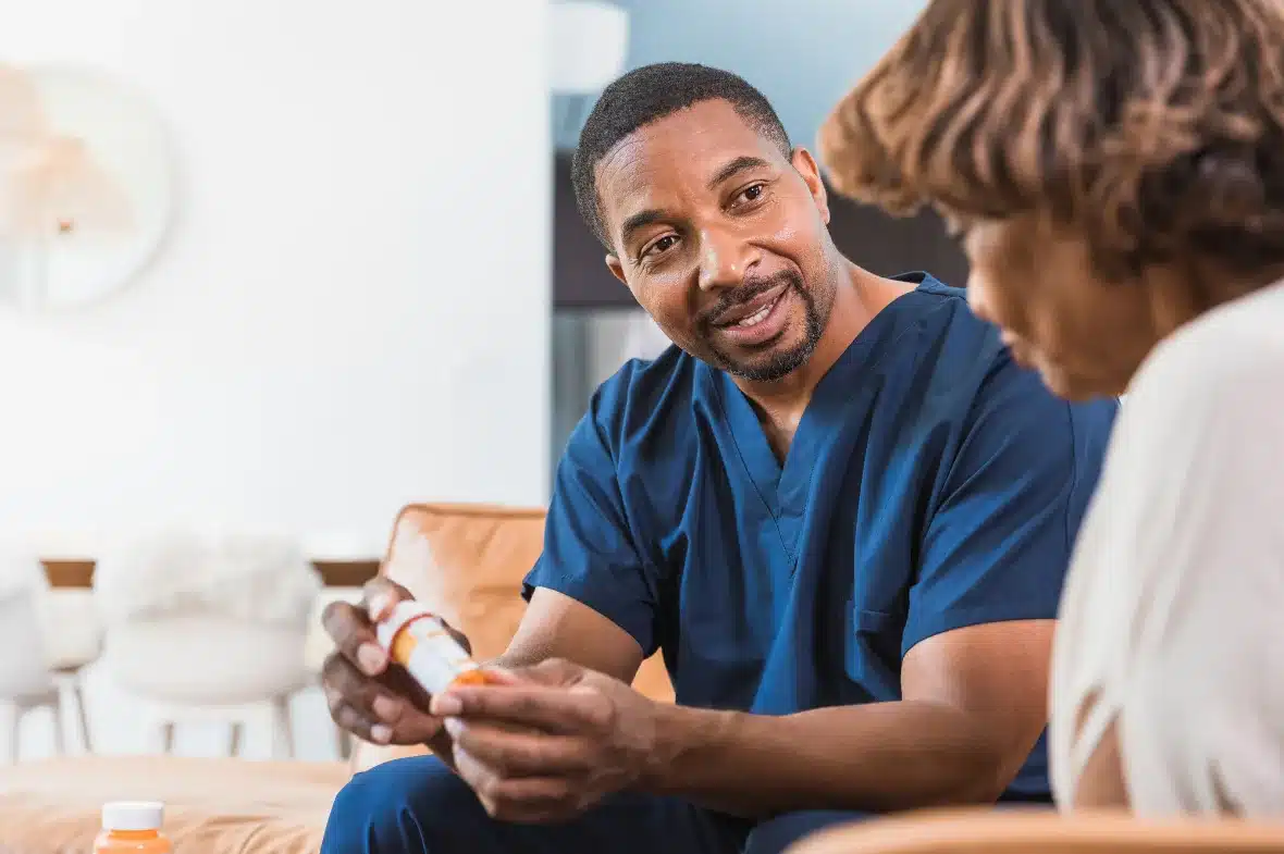 A healthcare professional, in blue scrubs, warmly explains a medication bottle to a patient. The setting is a cozy, well-lit room, conveying trust and care.