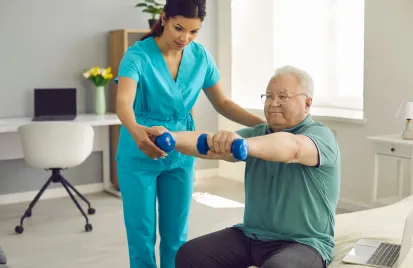 Elderly man exercising with dumbbells, assisted by a healthcare worker in scrubs. They are in a well-lit room with a desk and flowers, creating a supportive atmosphere.
