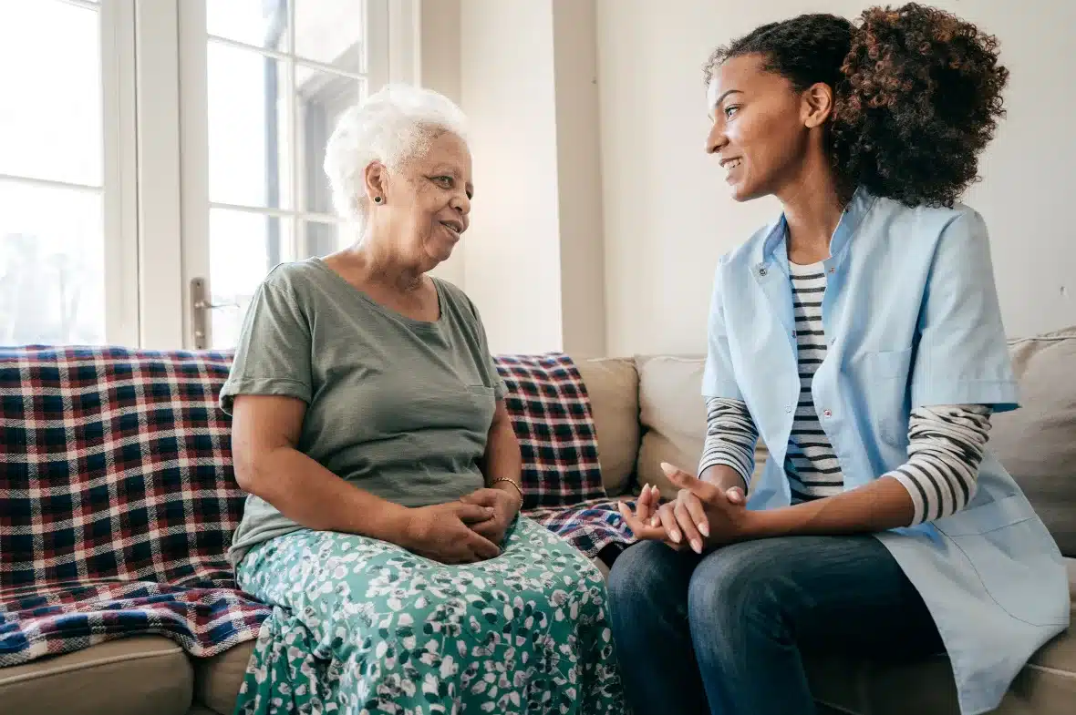 A caregiver in a blue uniform sits with an elderly woman on a couch with a checked blanket, sharing a warm, engaged conversation in a well-lit room.