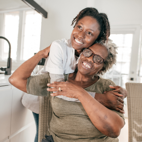 A joyful older woman with glasses sits in a kitchen, embraced from behind by a smiling younger woman. Bright light filters through large windows, creating a warm atmosphere.
