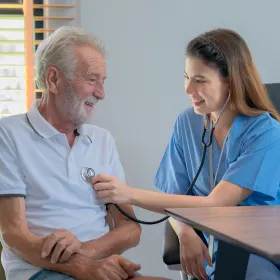 A nurse in blue scrubs smiles while using a stethoscope on an elderly man's chest. The room is bright, conveying a warm, caring atmosphere.