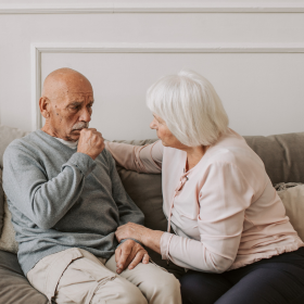 An elderly man sits on a sofa smiling at a nurse in a white uniform who is gently touching his shoulder. The setting is warm and supportive.