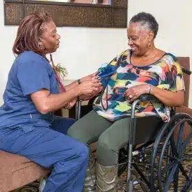 A nurse in blue scrubs checks the blood pressure of a smiling woman sitting in a wheelchair. The setting is warm and reassuring, highlighting care.