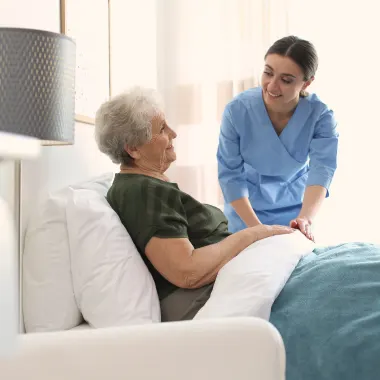 A nurse in blue scrubs gently assists an elderly woman sitting on a bed, creating a warm, caring atmosphere in a bright, softly-lit room.