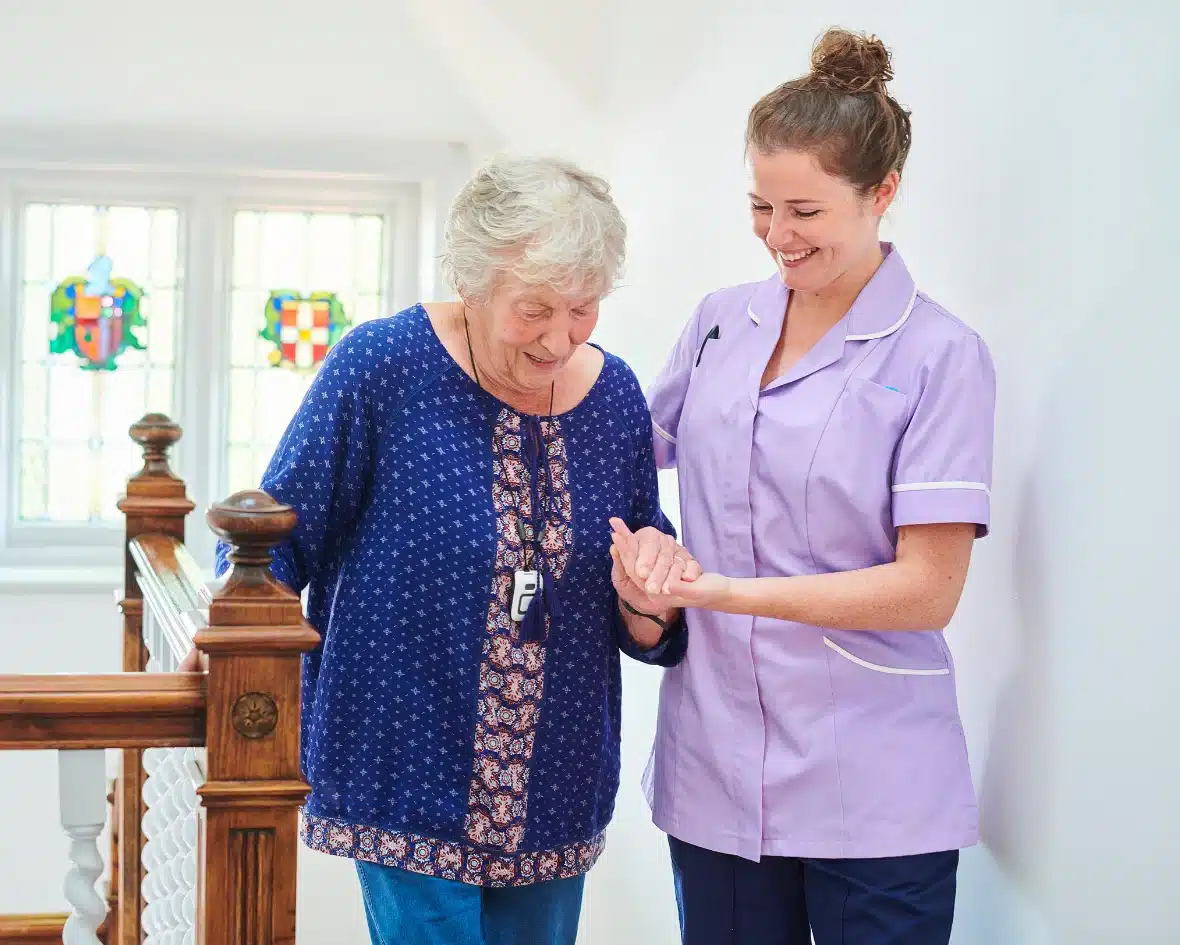 A caregiver in a purple uniform assists an elderly woman down a staircase, smiling. Bright stained glass windows add warmth to the supportive scene.