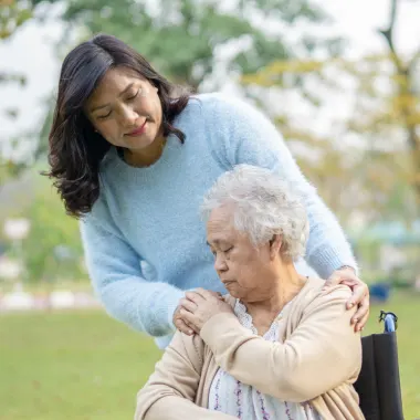 A woman in a blue sweater gently comforts an elderly woman in a wheelchair outdoors. The setting is a peaceful park with greenery, conveying care and empathy.