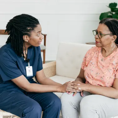 A healthcare professional in navy scrubs gently holds an older woman's hands on a sofa, conveying support and empathy in a calming environment.